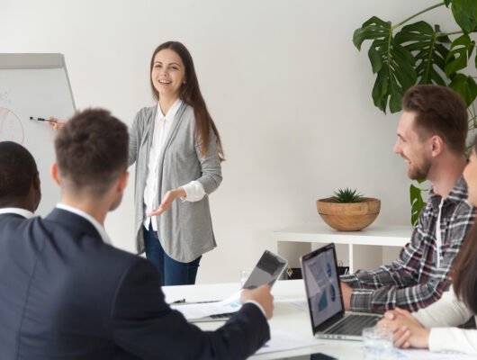 Smiling young employee or manager giving presentation working on flipchart in meeting room making business offer or reporting about project results, explaining new plan or idea to multi-ethnic team